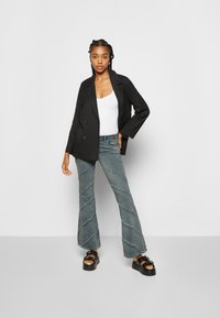 Blazer preto de dois botões, top branco justo, jeans azuis desbotados de perna larga com costura diagonal, e sandálias pretas chunky.