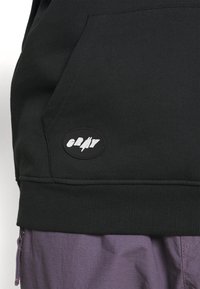 Černá mikina s kapucí vyrobená z bavlněné směsi, se žebrovaným lemem a přední kapsou. Ozdobená bílou kulatou nášivkou s logem na levé straně.