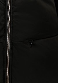 Chaqueta de plumas negra con una cremallera plateada brillante y un pequeño bolsillo con cremallera en el frente. La tela parece lisa y está aislada.