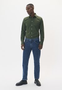 Zelena košulja s dugim rukavima, u kombinaciji s plavim trapericama. Crne cipele upotpunjuju odjevnu kombinaciju. Jednostavan, suvremen dizajn.