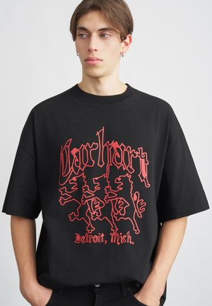 T-shirt nera in cotone con un audace design grafico rosso che presenta leoni stilizzati e il testo "Carhartt". Maniche corte, vestibilità comoda, colletto girocollo.