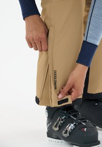 Beige waterdichte skibroek met zijritsen en klittenbandsluitingen, met het "W-PRO 10000" logo goed zichtbaar. Donkere skischchoenen zichtbaar daaronder.