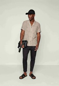 Man wearing beige short-sleeve button-up shirt, black cropped pants, black slides, and black cap, holding a black shoulder bag.