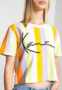 Nainen, joka on pukeutunut lyhythenkaiseen, vaaleanpunaisilla, keltaisilla, valkoisilla ja oransseilla pystysuorilla raidoilla varustettuun t-paitaan, jossa on mustaa käsin kirjoitettua tekstiä rinnassa.