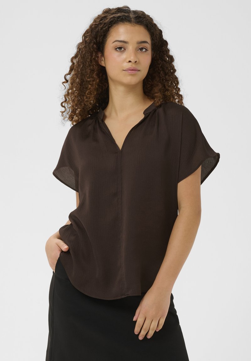 Dunkelbraune Bluse mit kurzen Ärmeln, V-Ausschnitt und lockerer Passform, aus strukturiertem Stoff mit dezentem Glanz, kombiniert mit einem schwarzen Rock.