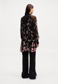 Haut long à manches transparentes noir à motif floral rose et vert, design superposé et ourlet à volants, associé à un pantalon à rayures noires.