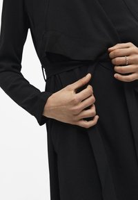 Vêtement noir à manches longues avec une taille nouée. Le tissu a une texture lisse. Les mains ajustent la ceinture, mettant en valeur un design simple.