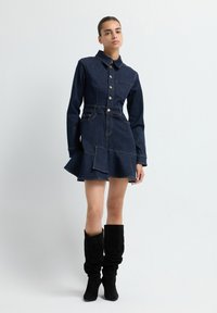 Robe chemise en denim bleu foncé avec un ourlet évasé, un devant à boutons, un col et des poches. Associée à des bottes noires à hauteur de genou et décontractées.