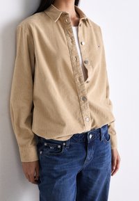 Tan corduroy shirt with a button-down collar, featuring silver buttons and a logo patch. Paired with dark blue denim jeans.