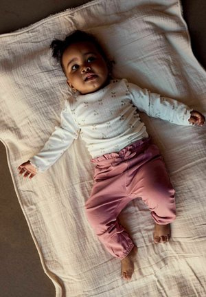 Niemowlę leżące na jasnym kocu, ubrane w białą bluzkę z długim rękawem z drobnymi wzorami oraz różowe spodnie, patrzące w górę z wyciągniętymi ramionami.