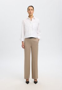 Camicia bianca con bottoni e tasca sul petto, abbinata a pantaloni beige a gamba larga e décolleté neri a punta. Sfondo semplice.