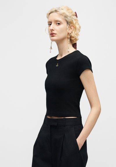 Ξανθιά γυναίκα με κοντά σγουρά μαλλιά που φοράει μαύρο κοντό τοπ, μαύρο παντελόνι ψηλής μέσης και μακριά κρεμαστά σκουλαρίκια με μαργαριτάρια.