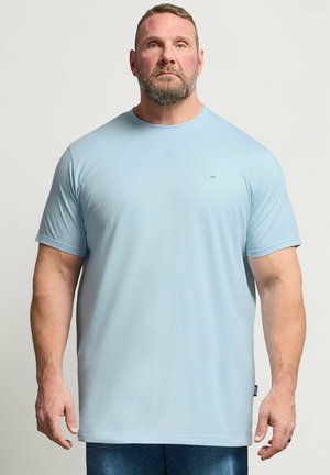 Mężczyzna z brodą, ubrany w jasnoniebieską koszulkę i dżinsy, stoi na tle jednolitego jasnego tła, zwrócony przodem z neutralnym wyrazem twarzy.