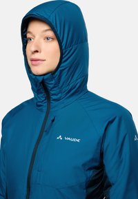 Chaqueta acolchada en color verde azulado con capucha, que presenta un cierre delantero de cremallera y un logo en el pecho. El material parece suave y ligero.