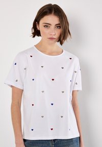 Hvid kortærmet t-shirt med spredte broderede hjerter i rød, blå og sølv. Rund halsudskæring med en afslappet pasform.