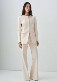 Traje beige a medida que presenta una chaqueta estructurada con escote redondo, pantalones acampanados y bolsillos laterales, confeccionado en una tela suave.