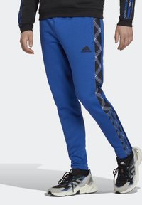 Blå atletiska joggingbyxor med en svart och blå rutmönstrad sidopanel, med en svart adidas-logotyp. Tillverkade av mjukt, stretchigt material.