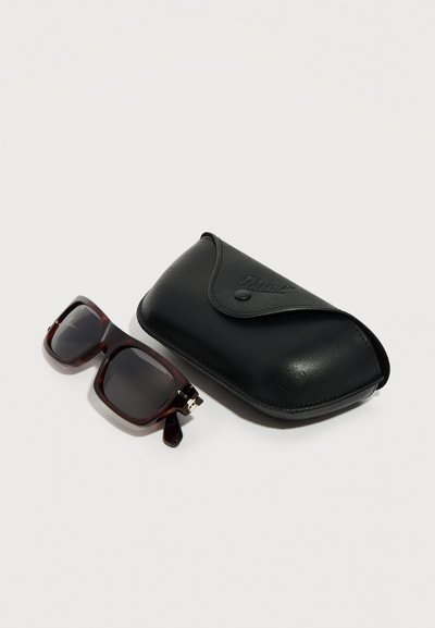 Γυαλιά ηλίου με καφέ καμβά και σκούρους φακούς δίπλα σε μαύρη σκληρή θήκη από δέρμα με κούμπωμα κουμπί σε λευκό φόντο.