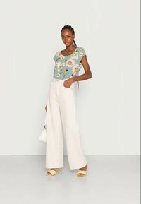 Top verde de manga curta com padrão floral, combinado com calças largas em bege claro e sandálias de salto bloco amarelas. Bolsa branca na mão.