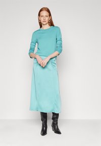Un haut à manches longues turquoise associé à une jupe longue en satin assortie. Le mannequin porte des bottes noires à talons hauts montant jusqu'aux genoux. Texture lisse dans l'ensemble.