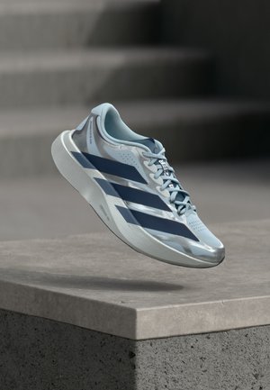 Chaussure de course bleu clair et argent avec des rayures bleu foncé flottant au-dessus d’un rebord en béton avec des marches floues en arrière-plan.