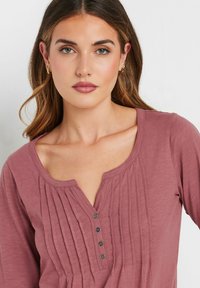Maglia a maniche lunghe color malva con scollatura a girocollo, dettagli plissettati e cinque bottoni sul davanti. Tessuto morbido.