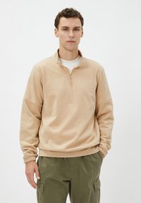 Sudadera de color beige con media cremallera en el cuello, puños elásticos y textura suave, combinada con pantalones cargo de color oliva.
