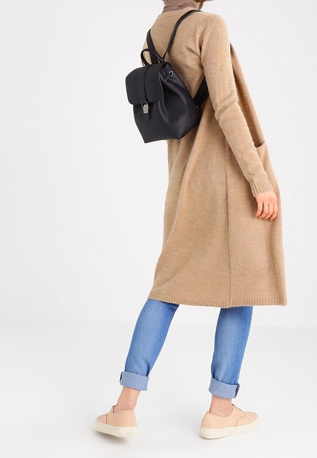 Vrouw draagt een beige lange cardigan, blauwe opgerolde spijkerbroek, beige sneakers en een kleine zwarte leren rugzak tegen een effen achtergrond.