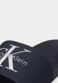 Μαύρη σαγιονάρα με λευκό κεντημένο λογότυπο Calvin Klein "CK" στο λουράκι, εμφανιζόμενη σε κοντινό πλάνο σε γκρι φόντο.