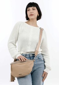 Bolso cruzado de cuero beige con cierre de cremallera y detalle de borlas. La correa es ajustable y cuenta con una hebilla de metal.
