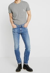 Camiseta de algodón gris con bolsillo y logo, combinada con jeans de mezclilla azules. El modelo lleva botas negras hasta el tobillo, de pie contra un fondo liso.