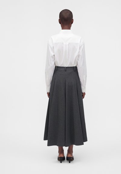 Άτομο που στέκεται με πλάτη, φοράει λευκό πουκάμισο με μακριά μανίκια, γκρι πλισέ midi φούστα και μαύρα παπούτσια με τακούνι σε απλό φόντο.