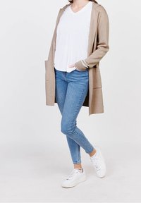 Person som bär en beige öppen cardigan, vit T-shirt, blå smala jeans och vita sneakers, stående med ett ben korsat framför det andra.