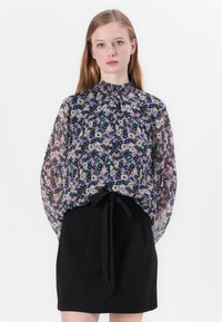 Blomstrete bluse med høy, rynket hals, gjennomsiktig stoff med rosa, hvite og lilla blomster, kombinert med et svart skjørt knyttet i livet.