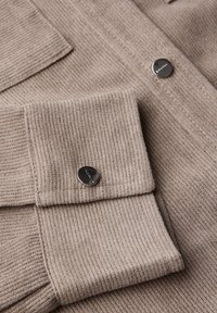 Beiger, gerippter Stoff am Hemdmanschetten und der Tasche mit silbernen Druckknöpfen, die die Textur und Details des Kleidungsstücks hervorheben.