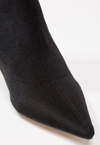 Gros plan d’un bout pointu de chaussure en tissu noir avec des coutures visibles sur fond blanc.