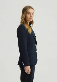 Navy blue tailored blazer made of smooth fabric, featuring silver buttons, a notch lapel, and detailed cuffs. Structured fit with side pockets.