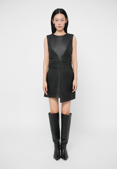Vestido de cuero negro con un diseño ajustado, tirantes sin mangas y dobladillo asimétrico, combinado con botas altas de tacón negro. Textura suave.