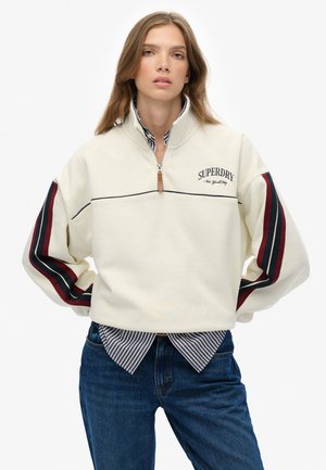 Sudadera de cremallera en el cuello de color crema, con rayas en azul marino y burdeos en las mangas, logo de SUPRDY y tejido texturizado. Combinada con una camisa a rayas y jeans.