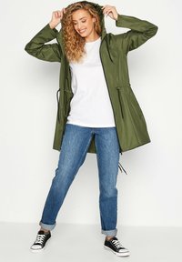Giacca a vento verde oliva con cappuccio e cordini, materiale leggero e polsi elasticizzati, abbinata a una maglietta bianca, jeans blu e scarpe da ginnastica nere.
