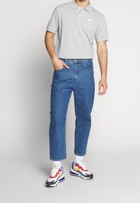 Grå bomullspolo med krage och vitt logo, kombinerad med blå denimjeans och flerfärgade sneakers med vit sula.