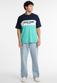 Īsām piedurknēm T-krekls ar tumši zilu, baltu un teļa krāsas paneļiem, uz kura ir "GUESS JEANS" logo. Kokvilnas audums, brīvs piegriezums, apaļa kakla izgriezums, ikdienas stils.
