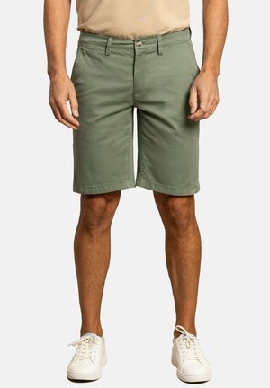 Pantalones cortos de carga verdes de algodón con un corte recto, que cuentan con un cierre de botón, presillas para cinturón y bolsillos laterales. Lleva puestos con zapatillas blancas.