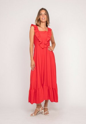 Femme portant une robe maxi rouge sans manches avec des volants le long du décolleté et de l'ourlet, debout avec la main sur la hanche et des sandales blanches à talons.