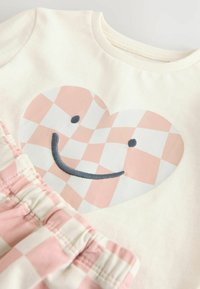 T-shirt crème clair avec un design de cœur à carreaux roses affichant un sourire et des coutures bleu marine ; short à carreaux roses et blancs avec une taille froncée.