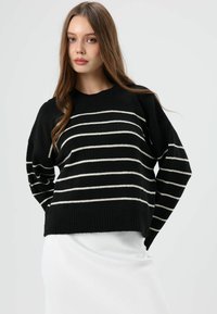 Pull en maille noire avec rayures horizontales blanches, ourlet et poignets côtelés, coupe décontractée, encolure ronde. Porté avec une jupe blanche.
