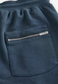 Pantaloni de trening bleumarin, realizati dintr-un material texturat, cu un buzunar posterior cu fermoar argintiu, accentuați de un tiv ribborat.