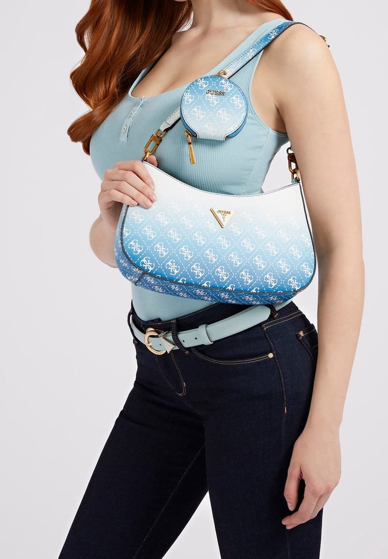 Femme portant un débardeur bleu clair avec un jean foncé et une ceinture bleu clair assortie, tenant un sac à main Guess bleu et blanc avec une petite pochette attachée.