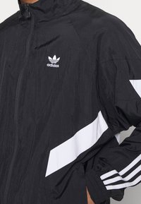 Svart idrottsjacka med dragkedja, vita detaljer och tre ränder på ärmarna. Materialet har en strukturerad yta.