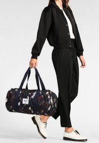 Granatowa torba wojskowa z kwiatowym i ptasim wzorem, solidnymi granatowymi uchwytami i białą naszywką z logo. Modelka ma na sobie czarną kurtkę i spodnie.